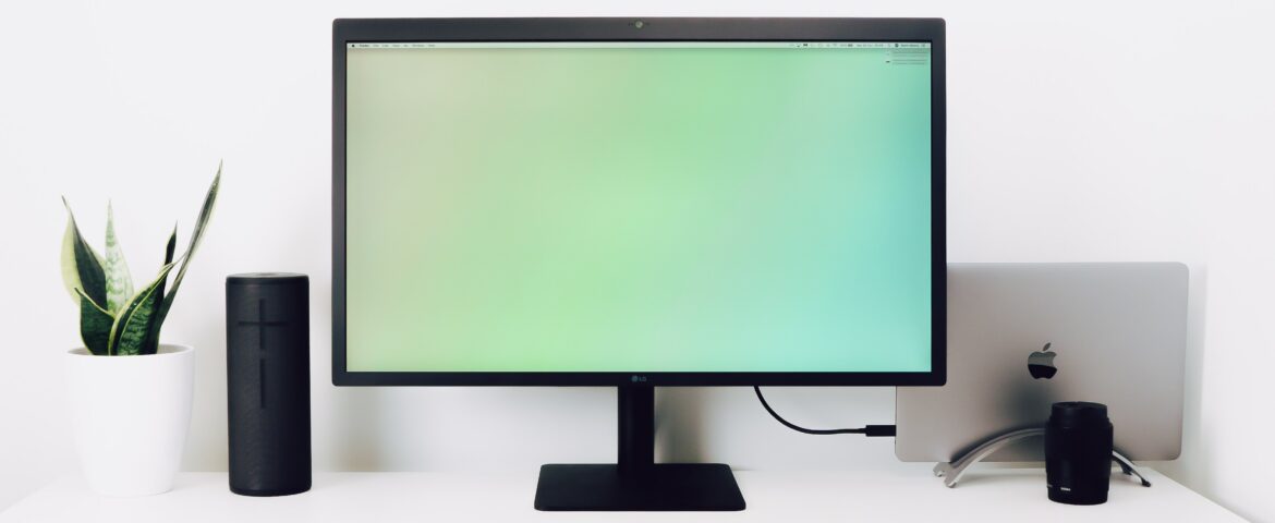 decorative image: a work desk with a big monitor, speakers, a keyboard and a mobile phone. In the back is a laptop stashed away and on the left you can see a plant.