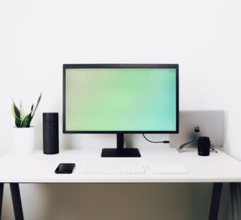 decorative image: a work desk with a big monitor, speakers, a keyboard and a mobile phone. In the back is a laptop stashed away and on the left you can see a plant.